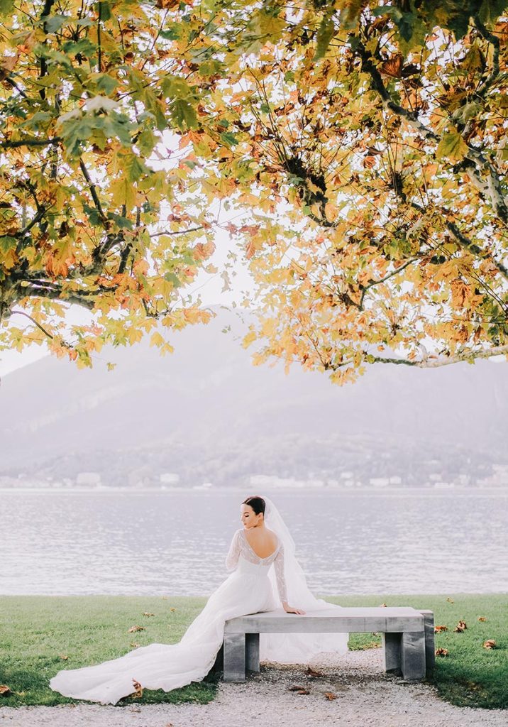 A bride in an elegant gown and veil sits on a bench beneath autumn leaves, with Lake Como and mountains in the background, reflecting Lake Como wedding styles