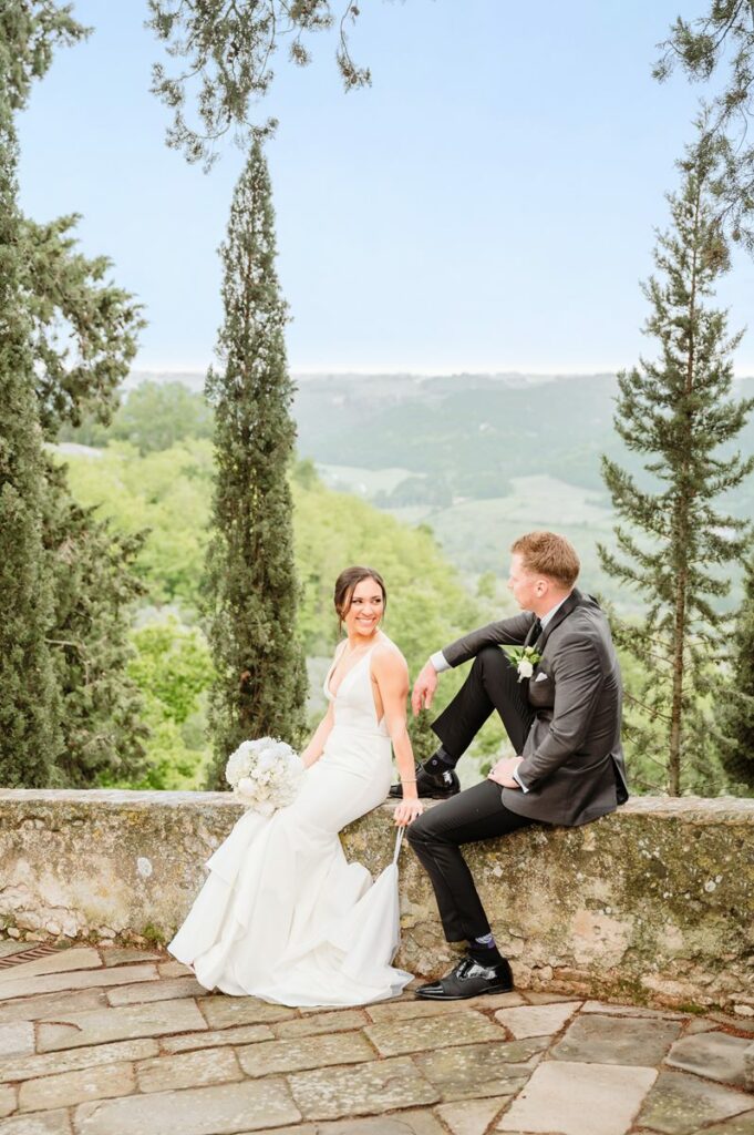 Bride and groom sitting on a stone wall surrounded by cypress trees, with rolling Tuscan hills in the background