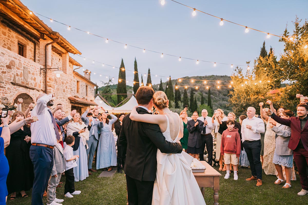 Bride and groom embracing as wedding guests raise their glasses in celebration at Villa San Crispolto, under glowing string lights