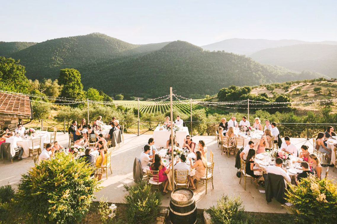 A charming outdoor wedding reception at a budget-friendly Tuscan venue, featuring round tables, string lights, and a view of vineyards and rolling hills.