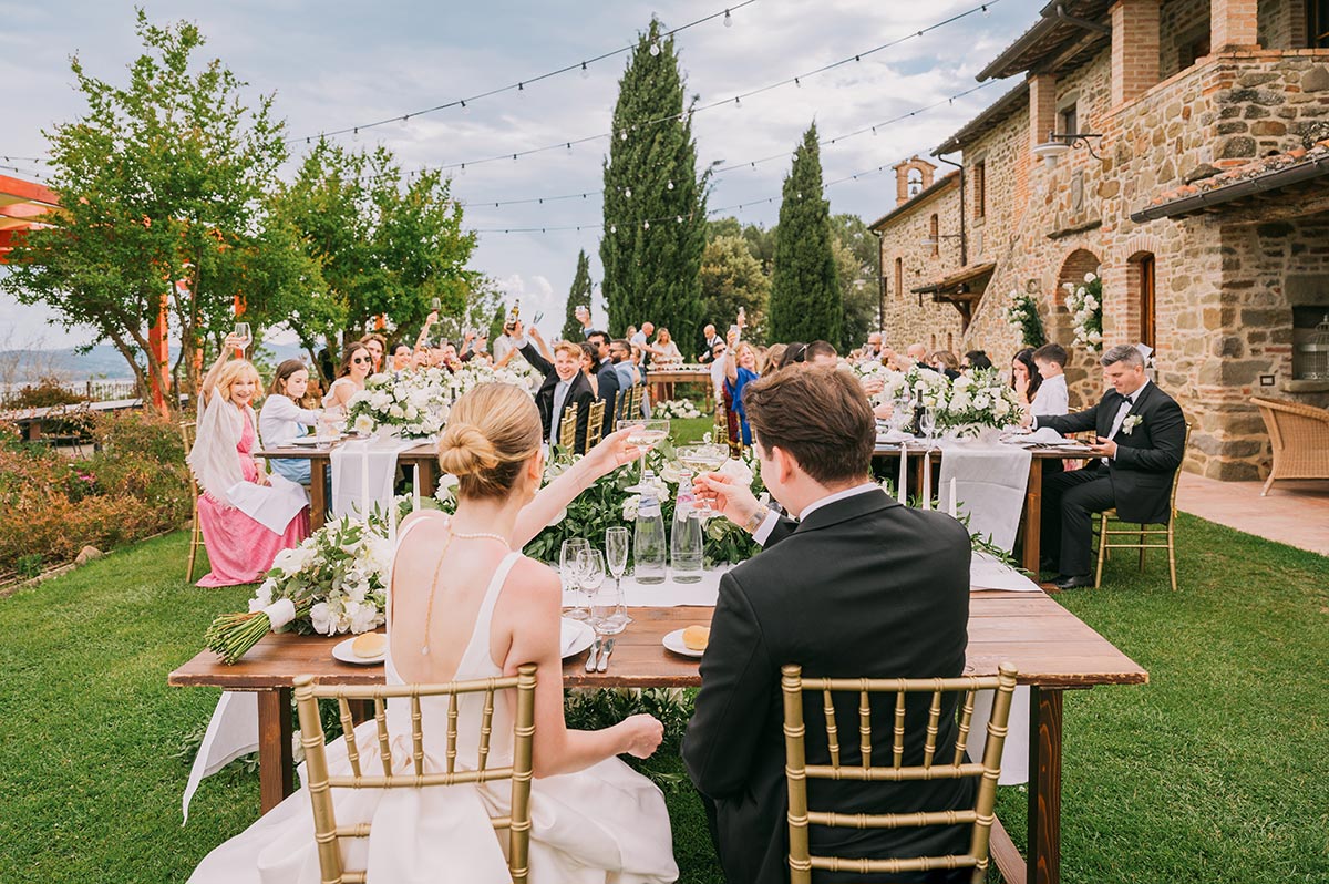 Newlywed couple raising glasses at an outdoor wedding reception in a rustic Tuscan villa garden with guests seated under string lights