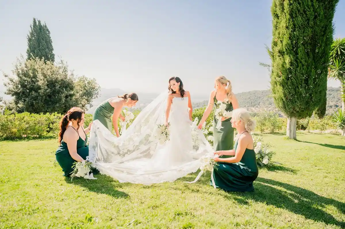 Alt: A bride in a flowing white gown with bridesmaids in green dresses adjusting her train, set against a sunny Tuscan countryside